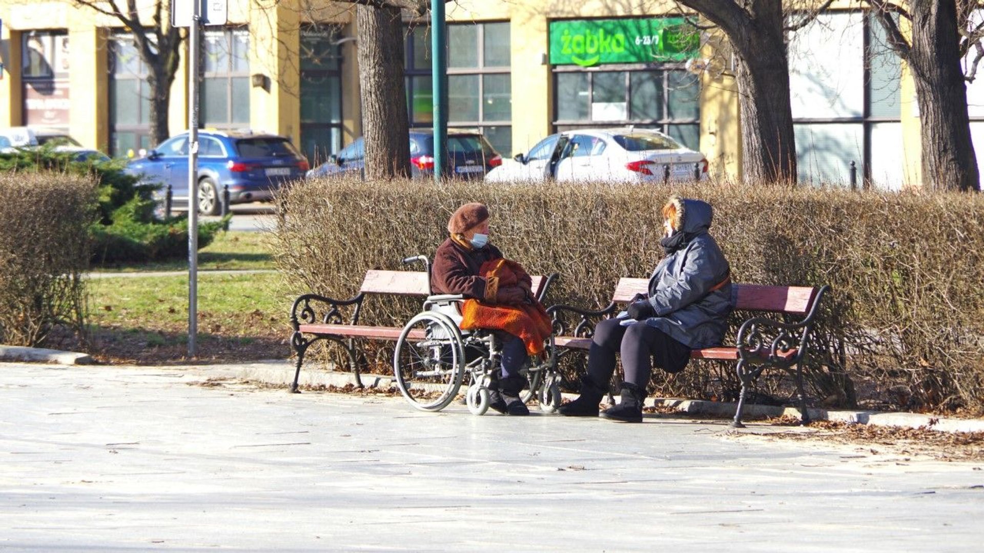 Dwie emerytki rozmawiają w maseczkach na ławce, jedna na wózku inwalidzkim