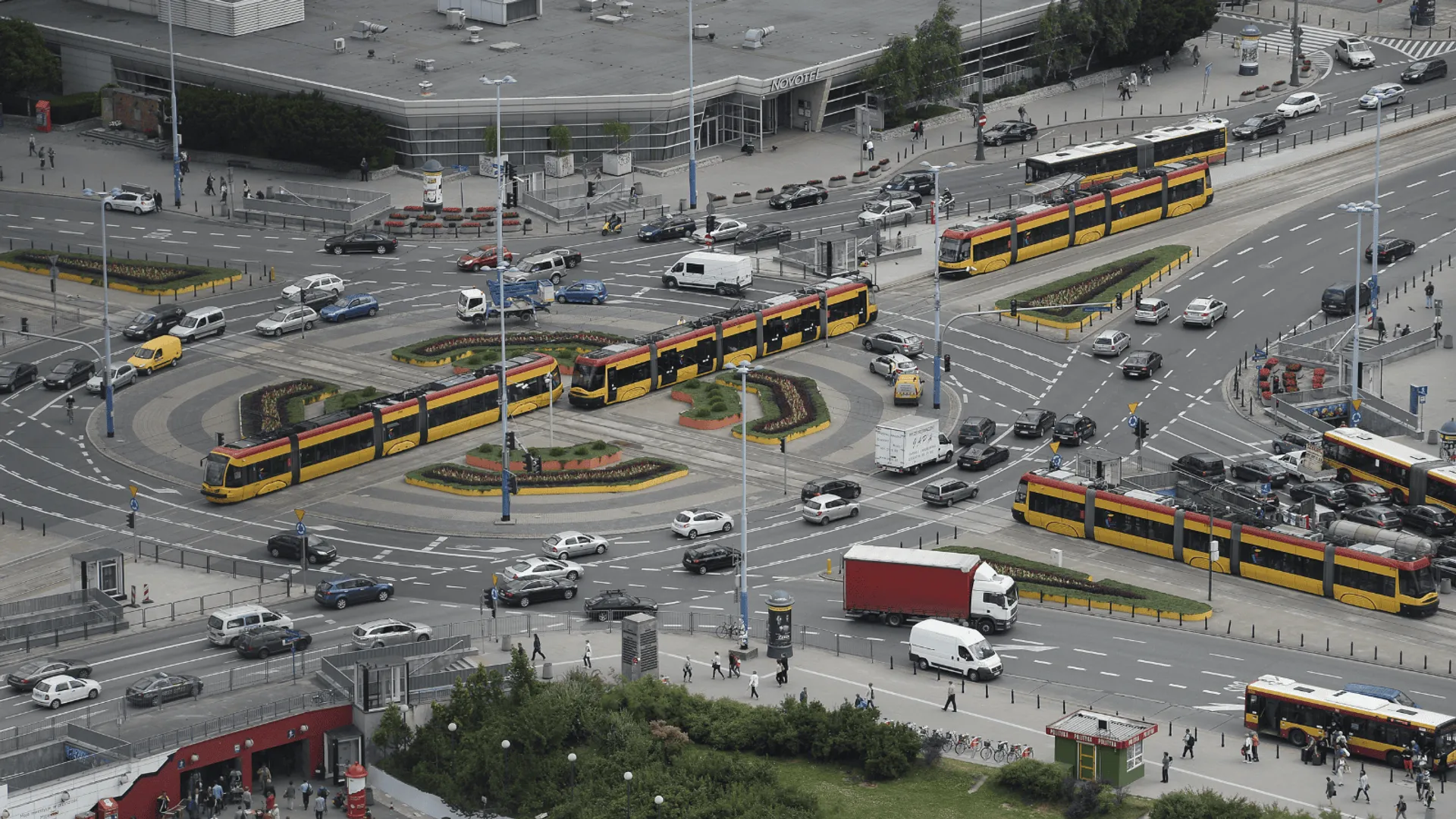 Widok z góry na rondo w centrum Warszawy z licznymi tramwajami, samochodami i pieszymi, przedstawiający intensywny ruch miejski