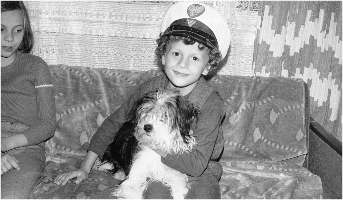 Chłopiec w milicyjnej czapce z psem na kolanach i dziewczynka siedzą na kanapie. 1977 r.  