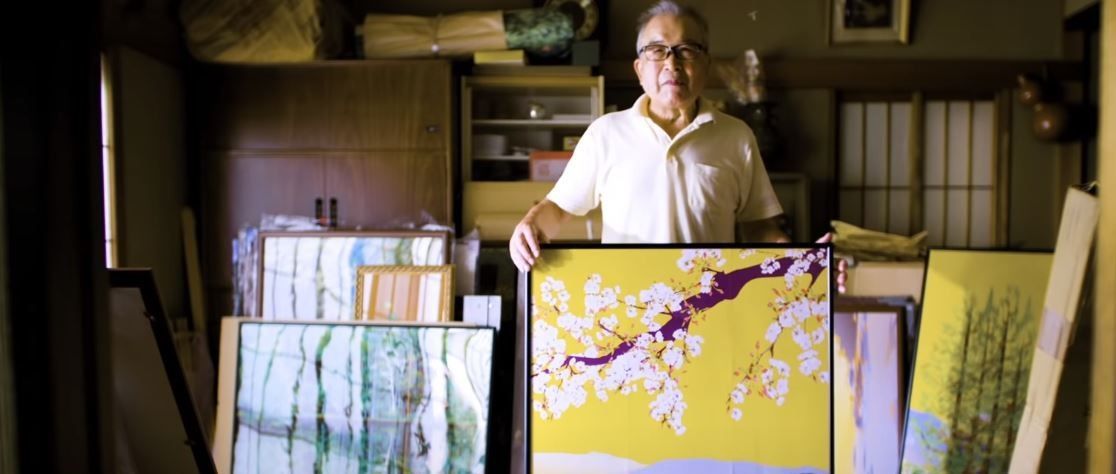 Tatsuo, 80-letni Japończyk prezentuje swój obraz tworzony w programie Microsoft Excel. Starszy mężczyzna trzyma obraz na środku pokoju. W tle inne obrazy.