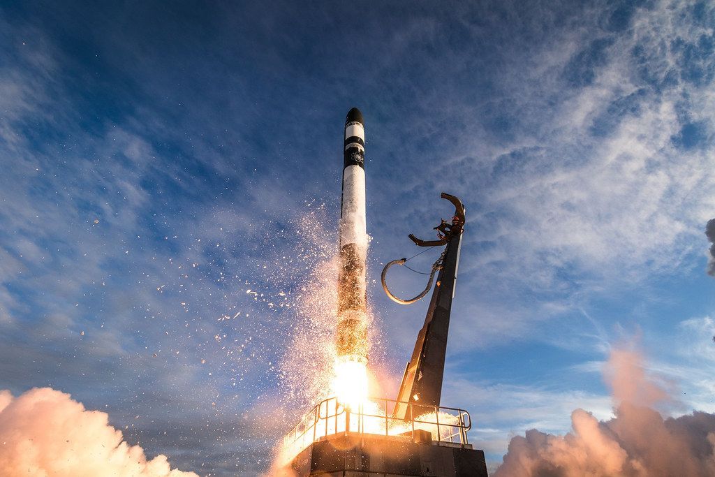 Rakieta Electron należąca do firmy Rocket Lab. Zdjęcie wykonane podczas startu, ogień z dyszy paliwowej rakiety, kosmodrom, niebieskie niebo, lekkie zachmurzenie.