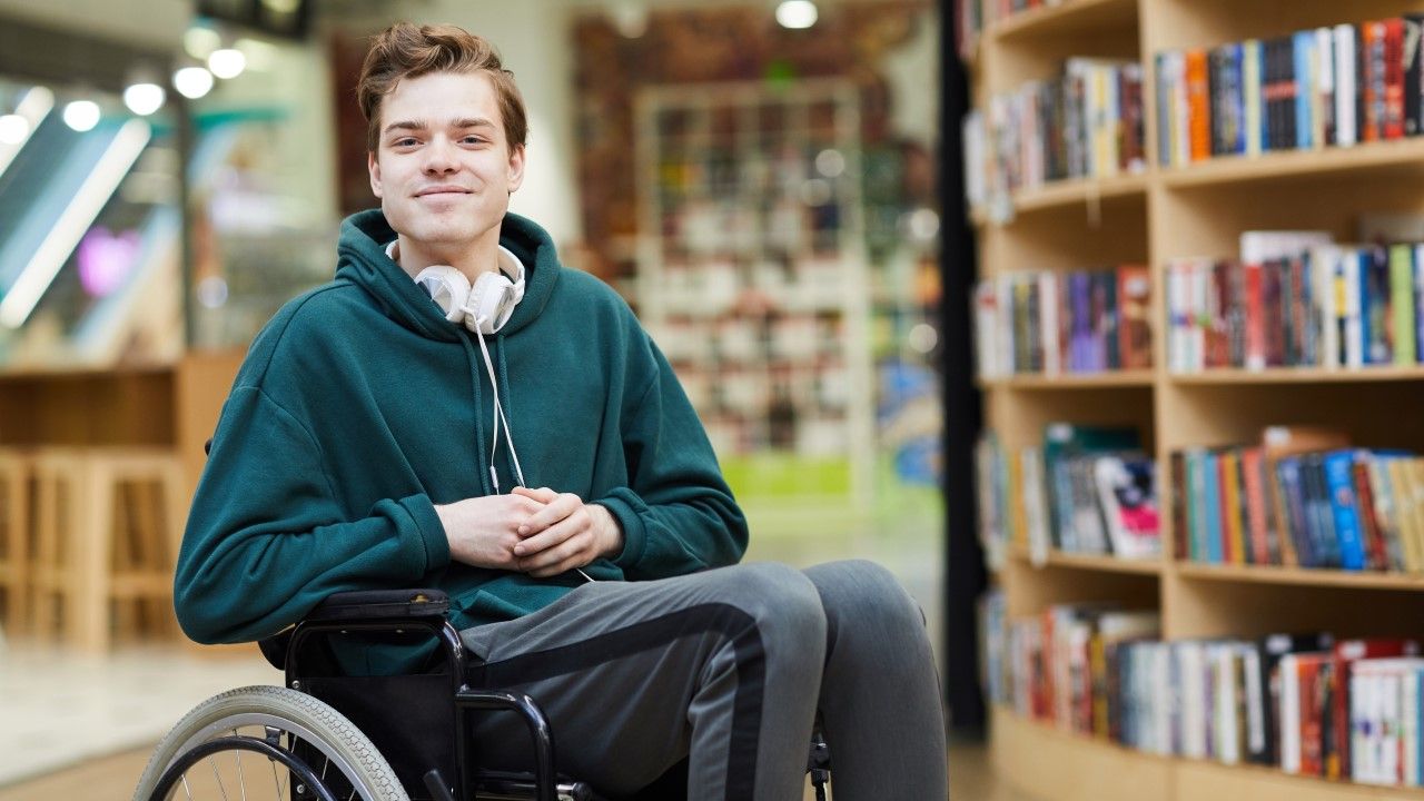 Uśmiechnięty chłopak na wózku inwalidzkim, w tle biblioteka