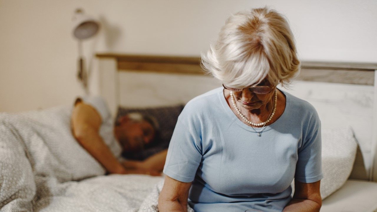 Starsza kobieta siedzi nocą na brzegu łóżka, obok śpi mąż