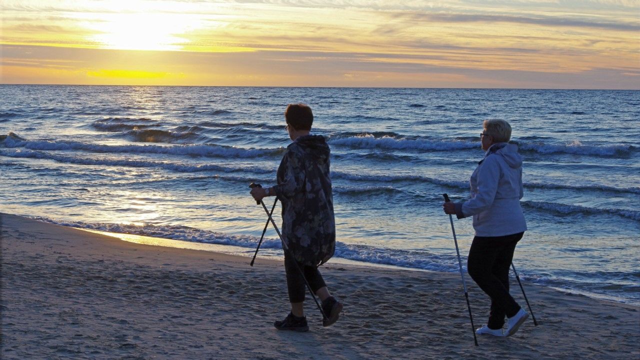 Dwie starsze panie spacerujące z kijkami nad morzem, w tle zachód słońca